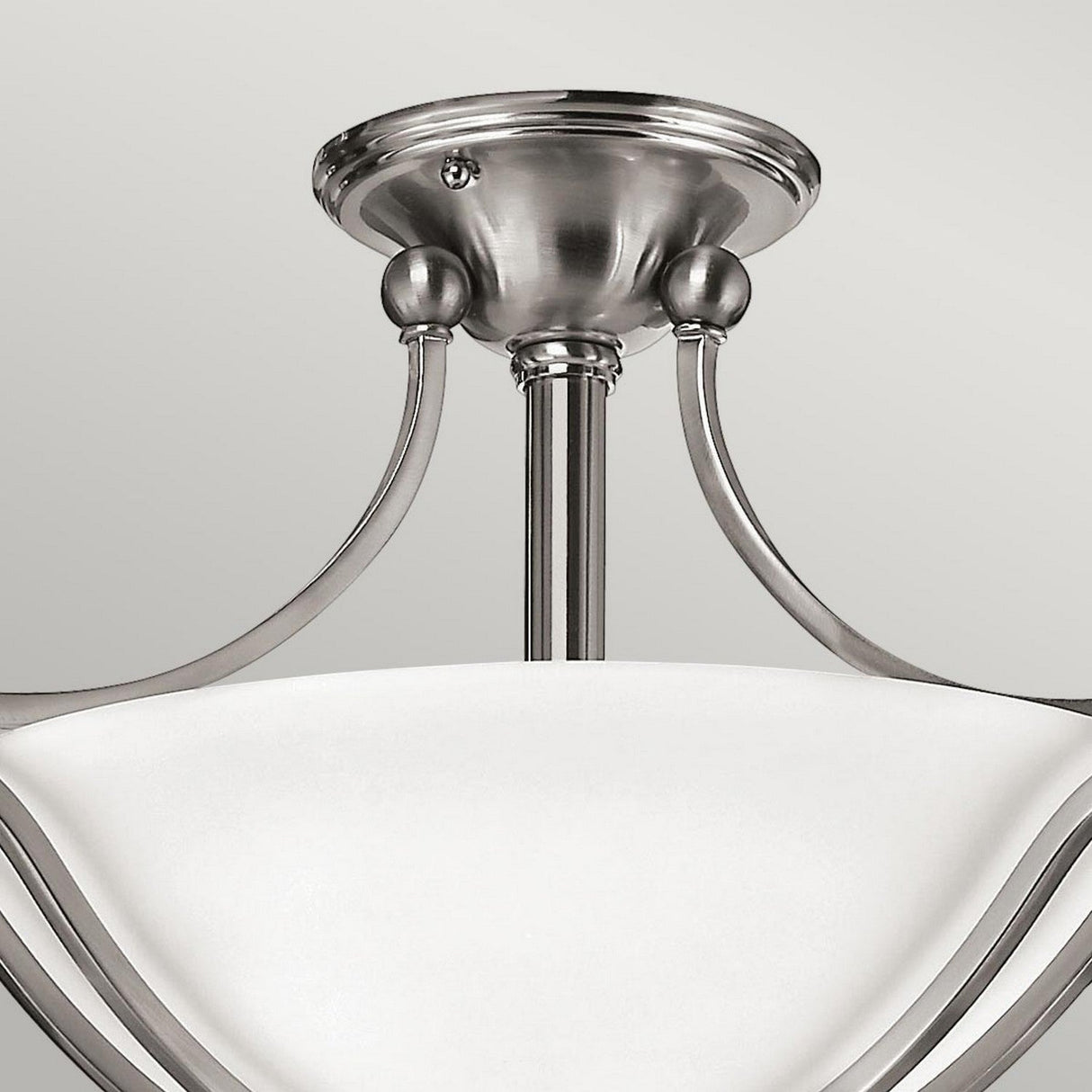 The Bolla 2 Light Semi Flush- Brushed Nickel fixture, ideal for sophisticated home decor, features a refined brushed nickel finish. Its smoothly curved white glass shade is elegantly upheld by three stylish metal arms extending from a central stem attached to a round ceiling mount.