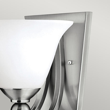 Close-up of the Bolla 1 Light Wall Light in brushed nickel, showcasing its bell-shaped glass shade and a brushed metal base with curved details. The sleek, contemporary design is an elegant decorative accent against a neutral backdrop.