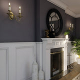 A stylish living room corner with New England style features a fireplace and a decorative mirror above it. The walls are half-paneled and painted gray. Two Cambridge 2 Light Wall Lights in burnished brass with optic glass shades add light, while potted plants provide a natural touch. White and black decor accents complete the scene.