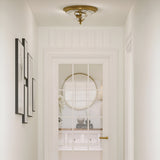 A well-lit hallway with white walls and framed artworks on the left exudes New England decor charm. The door, featuring glass panes, offers a view of a round mirror and books on a console table. Positioned above and enhancing the elegance of the setting is the Cambridge 2 Light Flush in Burnished Brass.