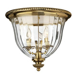 The Cambridge 3 Light Flush - Burnished Brass is a New England style ceiling fixture designed with clear glass panels and three candle-style bulbs. It boasts an elegant, vintage-inspired appearance, showcasing a decorative brass rim and intricate detailing at its base.