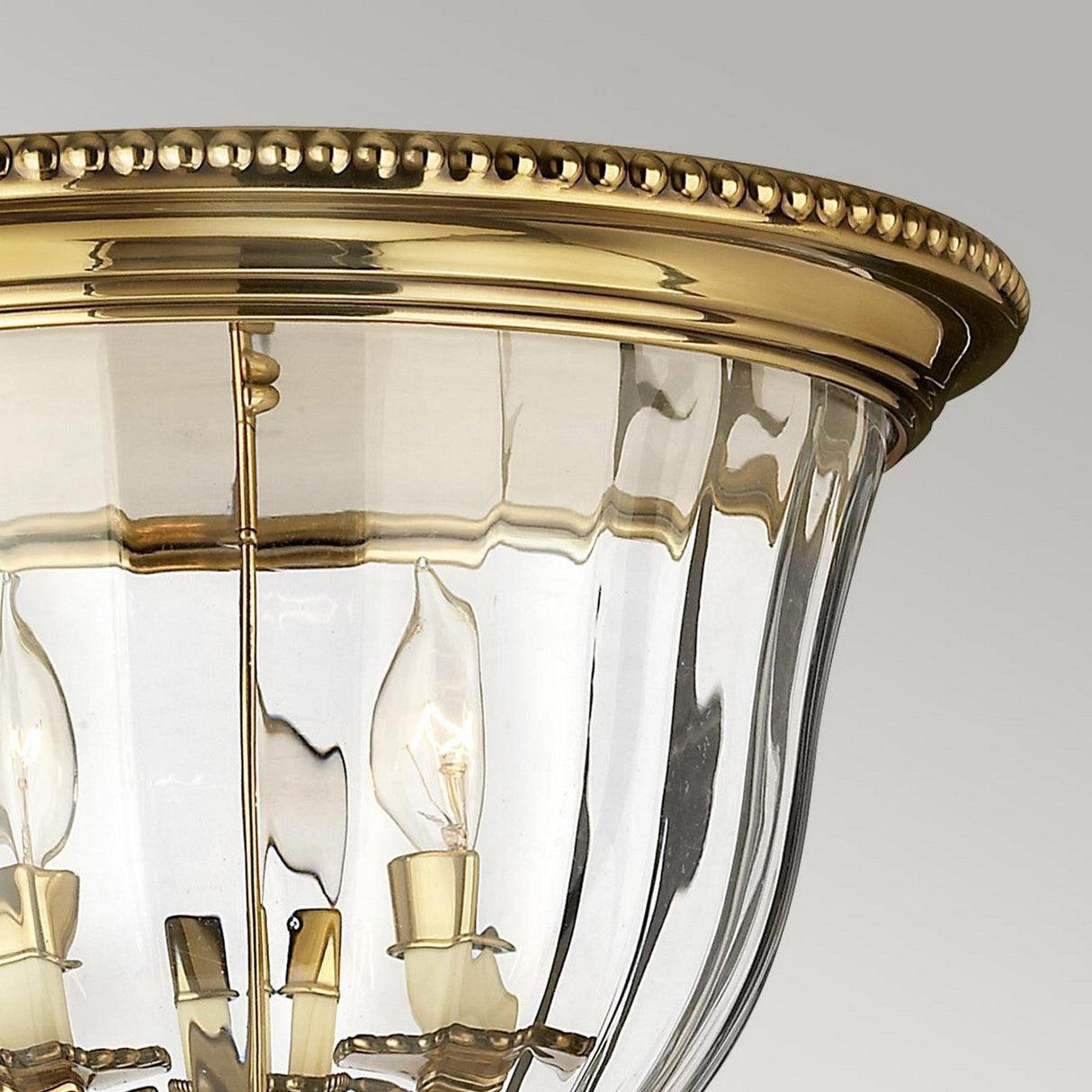 A close-up of the Cambridge 3 Light Flush - Burnished Brass reveals an ornate ceiling fixture with a clear glass shade. Its elegantly carved rim houses multiple candle-shaped bulbs, capturing the classic New England style against a soft gray background.