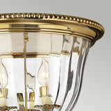 A close-up of the Cambridge 3 Light Flush - Burnished Brass reveals an ornate ceiling fixture with a clear glass shade. Its elegantly carved rim houses multiple candle-shaped bulbs, capturing the classic New England style against a soft gray background.