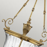 Close-up of a Cambridge 3 Light Chandelier - Burnished Brass. This elegant fixture, crafted from solid brass, features ornamental detailing and intricate metalwork with curved accents and a decorative finial. The glass enclosure stands out against the plain, neutral background.