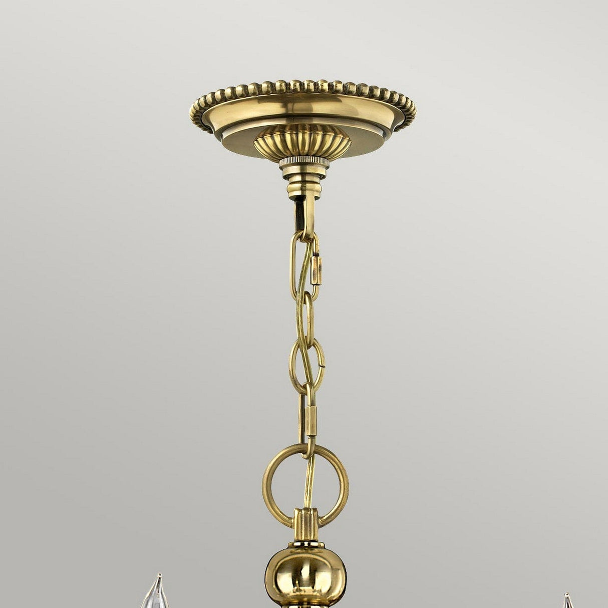 A close-up of a burnished brass ceiling mount showcases an elegant decorative design with a chain extending downwards. The round, polished links connect to a spherical ornament at the bottom. The plain, light gray background highlights the timeless allure reminiscent of the Cambridge 15 Light Chandelier - Burnished Brass.