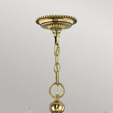 A close-up of a burnished brass ceiling mount showcases an elegant decorative design with a chain extending downwards. The round, polished links connect to a spherical ornament at the bottom. The plain, light gray background highlights the timeless allure reminiscent of the Cambridge 15 Light Chandelier - Burnished Brass.
