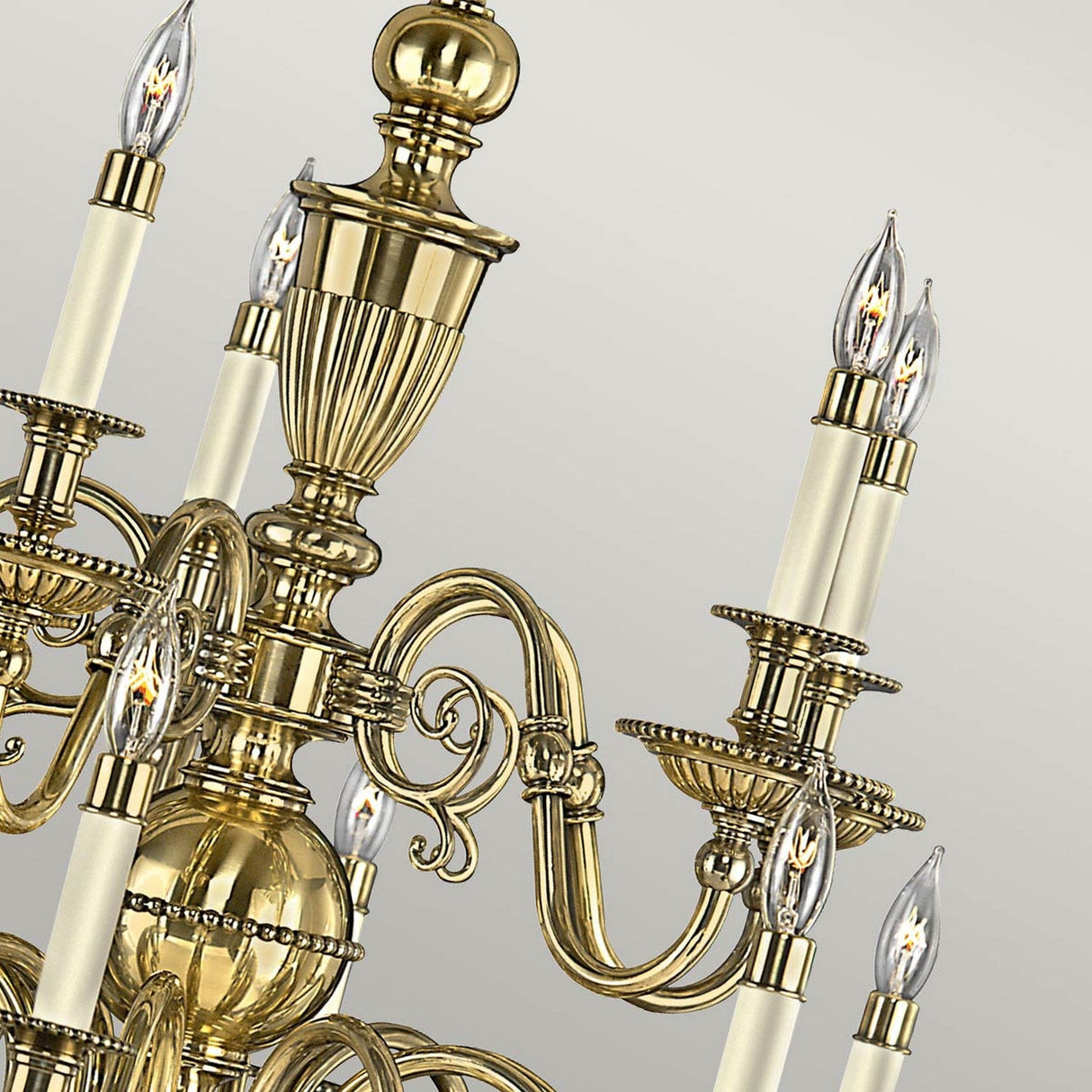 A detailed view of the Cambridge 15 Light Chandelier - Burnished Brass, highlighting its elegant scrollwork in a burnished brass finish. The chandelier includes numerous candle-style bulbs encased in clear glass, all set against a light gray background.