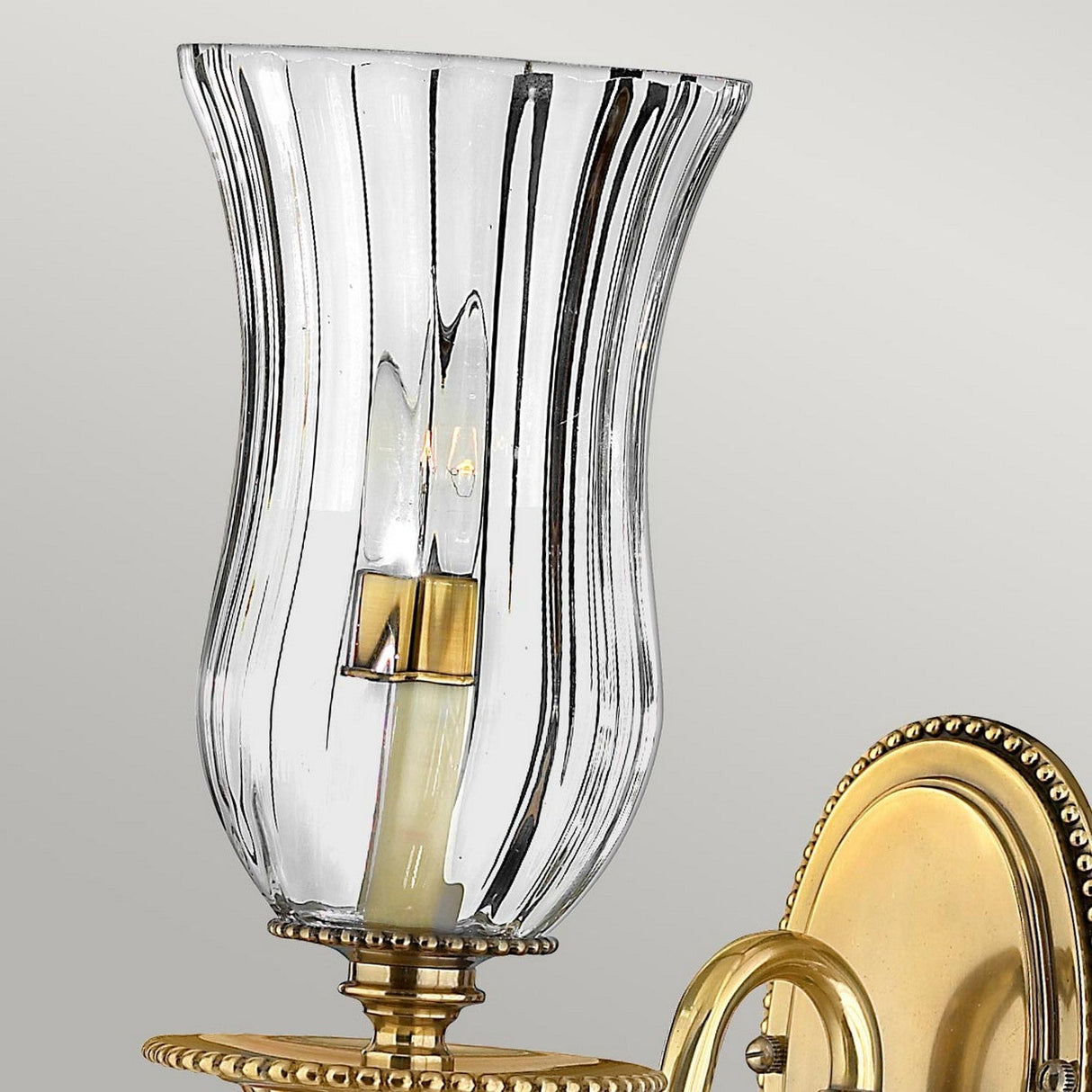 The Cambridge 1 Light Wall Light in burnished brass embodies classic New England elegance. Its clear, fluted glass shade showcases a visible bulb inside, complemented by a circular backplate adorned with intricate beaded detailing.