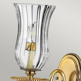 The Cambridge 1 Light Wall Light in burnished brass embodies classic New England elegance. Its clear, fluted glass shade showcases a visible bulb inside, complemented by a circular backplate adorned with intricate beaded detailing.