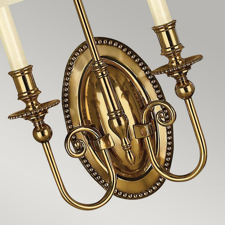 A close-up of the Cambridge 2 Light Wall Light in burnished brass showcases two candle-style fixtures. The refined design features intricate detailing and a polished finish, mounted on an oval backplate with beaded edges, exuding an elegant New England style.