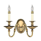 Introducing the Cambridge 2 Light Wall Light in burnished brass: This exquisite wall sconce showcases two candle-style lights with intricate detailing and a timeless, classic design. Each light boasts a slender white base, clear bulb tips, and optic glass shades, all elegantly mounted on ornate arms extending from an oval backplate.