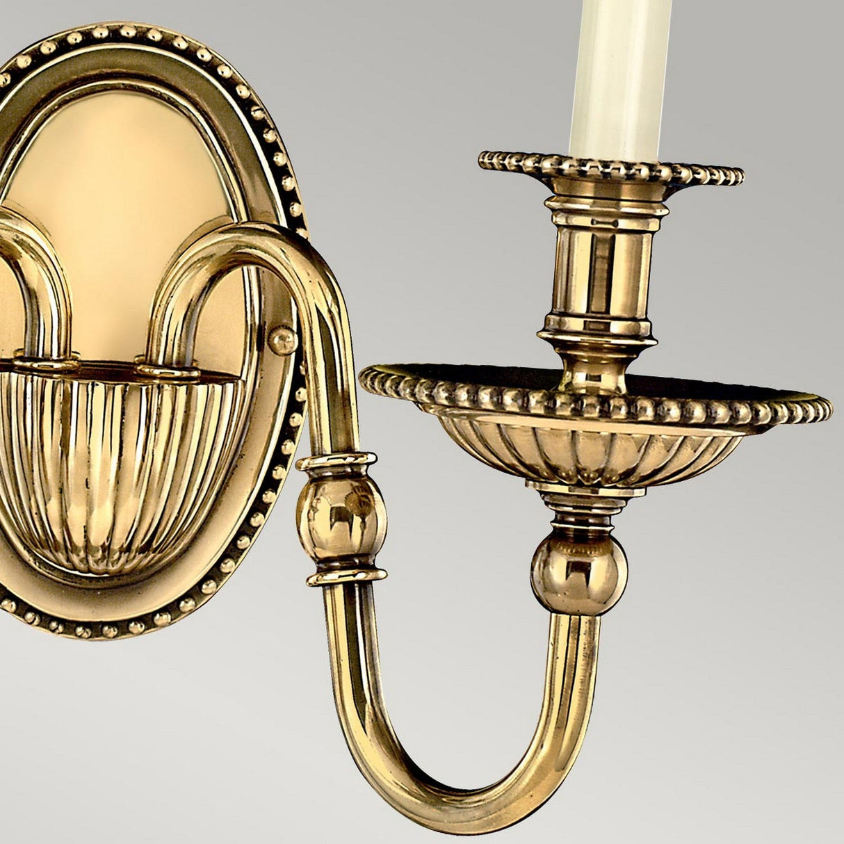 A close-up of the Cambridge 2 Light Wall Light in burnished brass showcases its ornate design, featuring intricate detailing and candle-like light fixtures. The New England style includes beaded edges, a curved arm with an oval backplate, set against a light gray background.