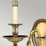 A close-up of the Cambridge 2 Light Wall Light in Burnished Brass, showcasing its candlestick-style design and intricate decorative detailing. Its smooth, gold finish shines against the plain gray backdrop, capturing the essence of New England style.