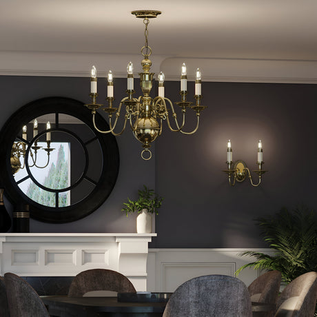 The sophisticated dining room is illuminated by a central Cambridge 6 Light Chandelier in burnished brass, featuring candle-like lights. A round table is surrounded by gray chairs, while a large mirror reflects the greenery. The dark walls are enhanced with matching candle-style wall sconces, also in burnished brass.