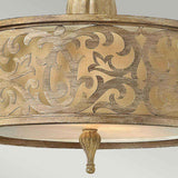 This image features a detailed view of a ceiling light fixture from the Carabel 2 Light Small Semi Flush collection in a brushed champagne finish. It is adorned with a gold-toned cylindrical shade showcasing intricate swirling patterns, and includes an elegant decorative finial hanging from its base.