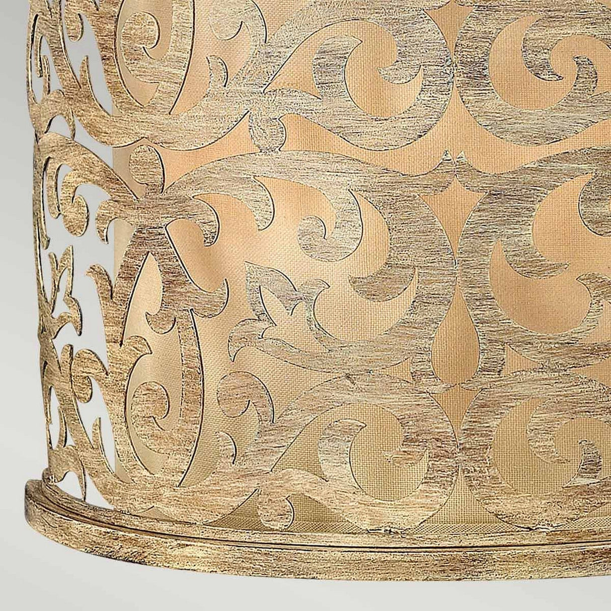 Close-up of the Carabel 2 Light Wall Light - Brushed Champagne showcasing an ornate metal pattern with intricate swirling designs on its cylindrical structure. The brushed champagne finish against a warm beige fabric background accentuates the elegant craftsmanship of the pierced metalwork.