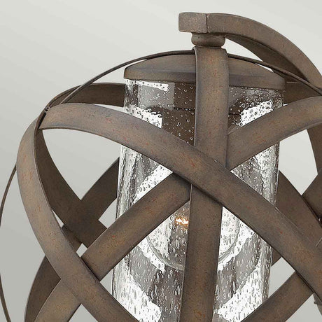 Introducing the Carson 1 Light Outdoor Small Wall Lantern in Vintage Iron: A glass cylinder envelops a bulb and is surrounded by gracefully curved metal bands, creating a spherical design. Its vintage iron finish lends an outdoor wall lantern a rustic, weathered charm.