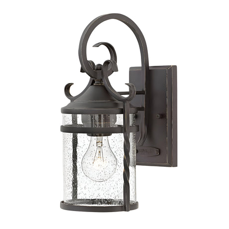 The Casa 1 Light Outdoor Wall Lantern - Black exudes rustic charm with its decorative design, made from black wrought iron and clear glass casing. It showcases a single visible light bulb inside, enhanced by a scroll motif on top and a rectangular mounting plate.