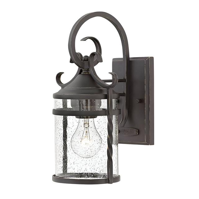 The Casa 1 Light Outdoor Wall Lantern - Black exudes rustic charm with its decorative design, made from black wrought iron and clear glass casing. It showcases a single visible light bulb inside, enhanced by a scroll motif on top and a rectangular mounting plate.