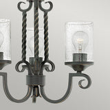 A close-up of the Casa 3 Light Chandelier - Black showcases its twisted wrought iron design and elegant seedy glass cylinder shades. Within the glass, two visible bulbs emit a warm glow, enhancing its charm. Set against a plain light gray background, the intricate details of this chandelier are beautifully highlighted.