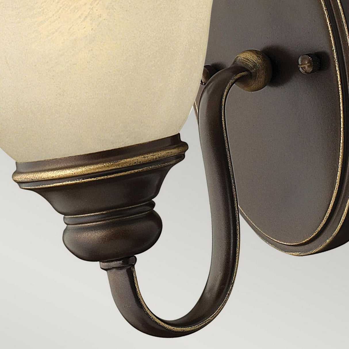 A detailed view of the Cello 1 Light Wall Light in an Antique Bronze finish, showcasing its ivory glass shade. The fixture includes a curved arm with intricate details and is mounted on a circular base, standing out against the neutral backdrop.