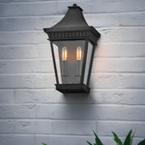 The Chapel Hill 2 Light Outdoor Half Wall Lantern in black is mounted on a white brick wall, its two candle-style bulbs aglow and green plant leaves visible at the bottom right, evoking classic carriage house charm.