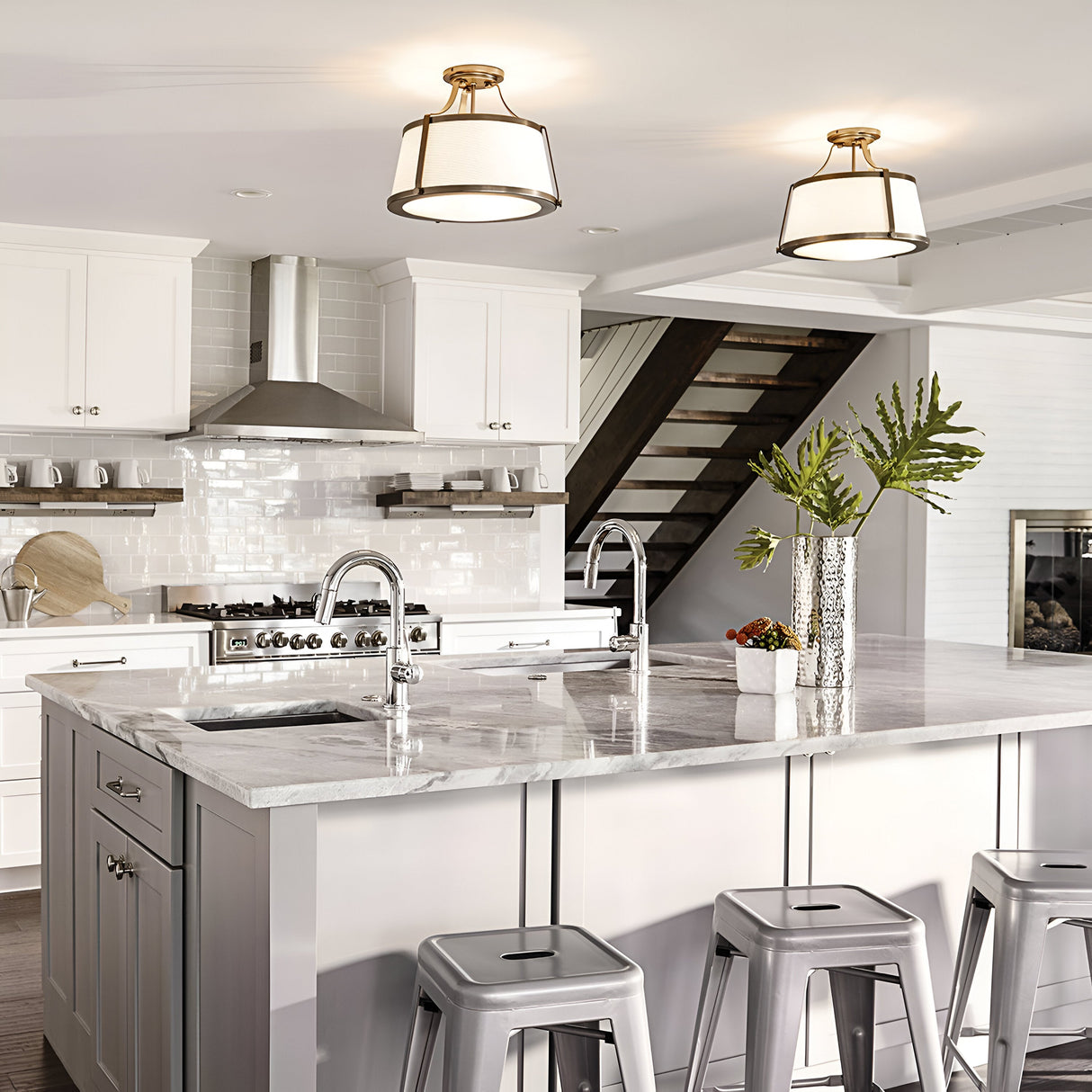 A modern kitchen featuring a marble island, gray stools, and stainless steel appliances is highlighted by white cabinets. Two pendant lights hang next to the Charlotte 3 Light Semi Flush fixture in brushed gold with a linen shade. Linen shade accents enhance the decorative plants on the countertop, while a staircase can be seen in the background.