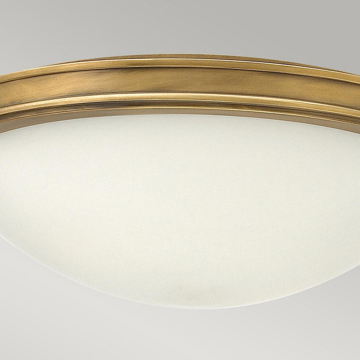 Introducing the Collier 3 Light Large Flush in Heritage Brass, a round ceiling-mounted fixture designed with a white frosted glass dome and elegantly framed by a Heritage Brass metallic rim, all set against a sophisticated gray backdrop.