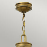 A close-up of the Collier 4 Light Chandelier in Heritage Brass showcases a ceiling light fixture with a chain connecting to its dome-shaped base. The chandelier's simple, elegant design features smooth curves and links, exuding mid-century charm against a plain grey background.