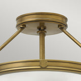 A close-up of the Collier 3 Light Semi Flush fixture in Heritage Brass showcases its circular design. With a sleek metallic finish and support from three angled rods, it presents an elegant and modern look against a simple backdrop.