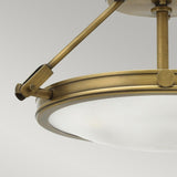 An up-close view of the Collier 3 Light Semi Flush in Heritage Brass showcases its opaque glass shade. This ceiling fixture features a circular design and a metal support arm, presented against a plain gray backdrop.