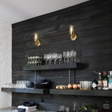 A cozy bar area exudes vintage charm with dark wooden shelves filled with various glassware, including wine glasses and mugs. Two Collier 1 Light Wall Lights in Heritage Brass provide warm lighting, while various bottles and decorative items are arranged on the counter below.