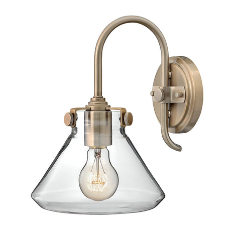 The Congress 1 Light Clear Glass Wall Light - Brushed Caramel features a curved arm and a clear, conical glass shade, displaying an exposed LED filament bulb. This fixture is mounted on a round base.