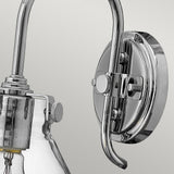 Close-up of the Congress 1 Light Clear Glass Wall Light in chrome finish, showcasing its round metal base and elegantly curved arm. The clear glass lampshade partially reveals the light bulb inside, while the chrome hardware elements offer a vintage touch to its modern design.