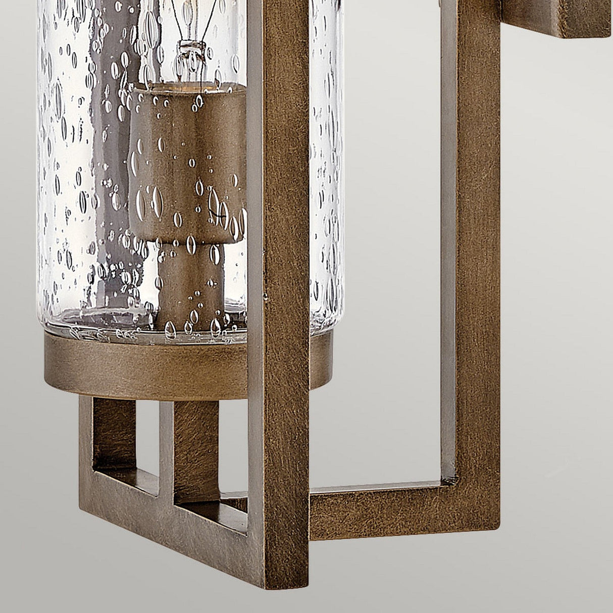 Close-up of the Cordillera 1 Light Outdoor Wall Light in burnished bronze, featuring a cylindrical clear glass cover with delicate bubbles that reveal a visible light bulb. This fixture embodies modern aesthetics against a plain, neutral background.