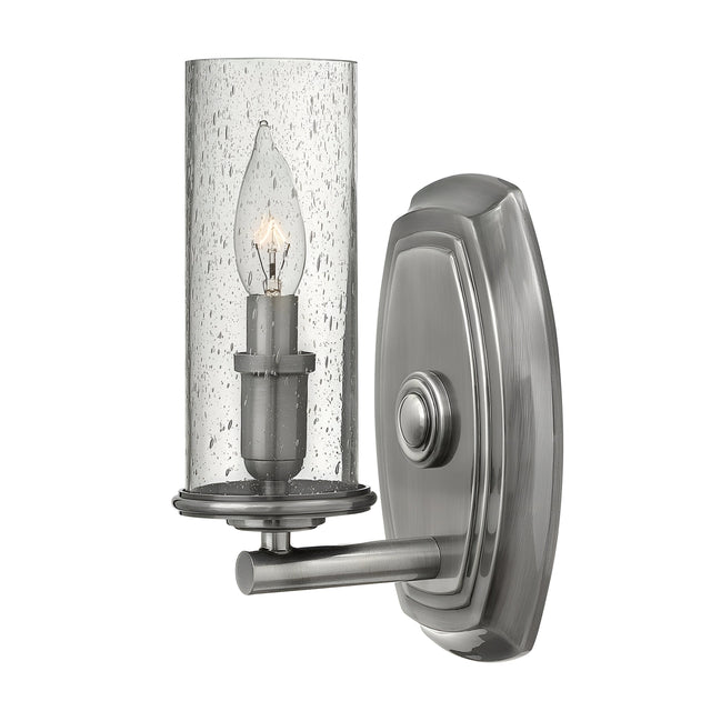 Introducing the Dakota 1 Light Wall Light - Nickel: a wall-mounted fixture featuring a cylindrical, transparent glass shade and candle-style bulb. Its metallic brushed nickel finish and sleek arm design provide transitional chic, perfectly complemented by an oval backplate for modern elegance.