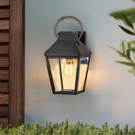 The Dawson 1 Light Outdoor Medium Wall Lantern in Black Oxide is mounted on a light wall by wooden siding, casting stylish illumination over green plants—an ideal choice for enhancing your patio lighting.