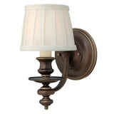 The Dunhill 1 Light Wall Light in a vintage style features a royal bronze finish and a cream-colored, pleated lampshade. Its transitional design includes ornate detailing and a curved arm, mounted on a circular base.