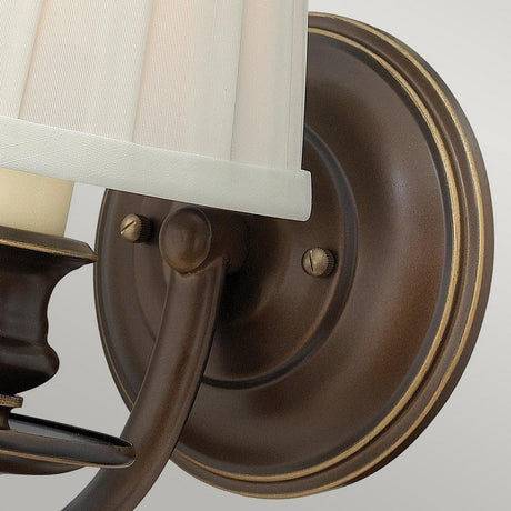 A close-up of the Dunhill 1 Light Wall Light - Bronze showcases its cylindrical, off-white fabric shade. The design includes a curved arm and circular backplate, embodying a transitional style with a classic, elegant appearance. It stands out beautifully against a light gray background.