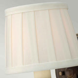 The Dunhill 1 Light Wall Light features a close-up view of its light beige lampshade, characterized by vertical fabric panels that embody a subtle transitional style. The shade is mounted on a dark, royal bronze fixture, partially visible against a light gray background, adding elegance to any wall lighting setting.