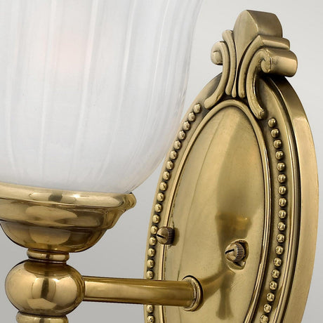 A close-up image of the Francoise 1 Light Wall Light in burnished brass, featuring a vintage design with an ornate oval base and a frosted ribbed glass lampshade. The base is adorned with decorative beading and top scrollwork, embodying a classic and elegant IP44 wall light.