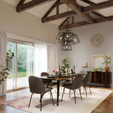In a contemporary dining room, a wooden table set for six is surrounded by dark chairs. The table is elegantly adorned with wine glasses and red napkins. Above it, a Fulton 7 Light Chandelier in Bronze & Brass with a minimalist design hangs from the beamed ceiling. Large windows offer stunning views of the lush green outdoor space.