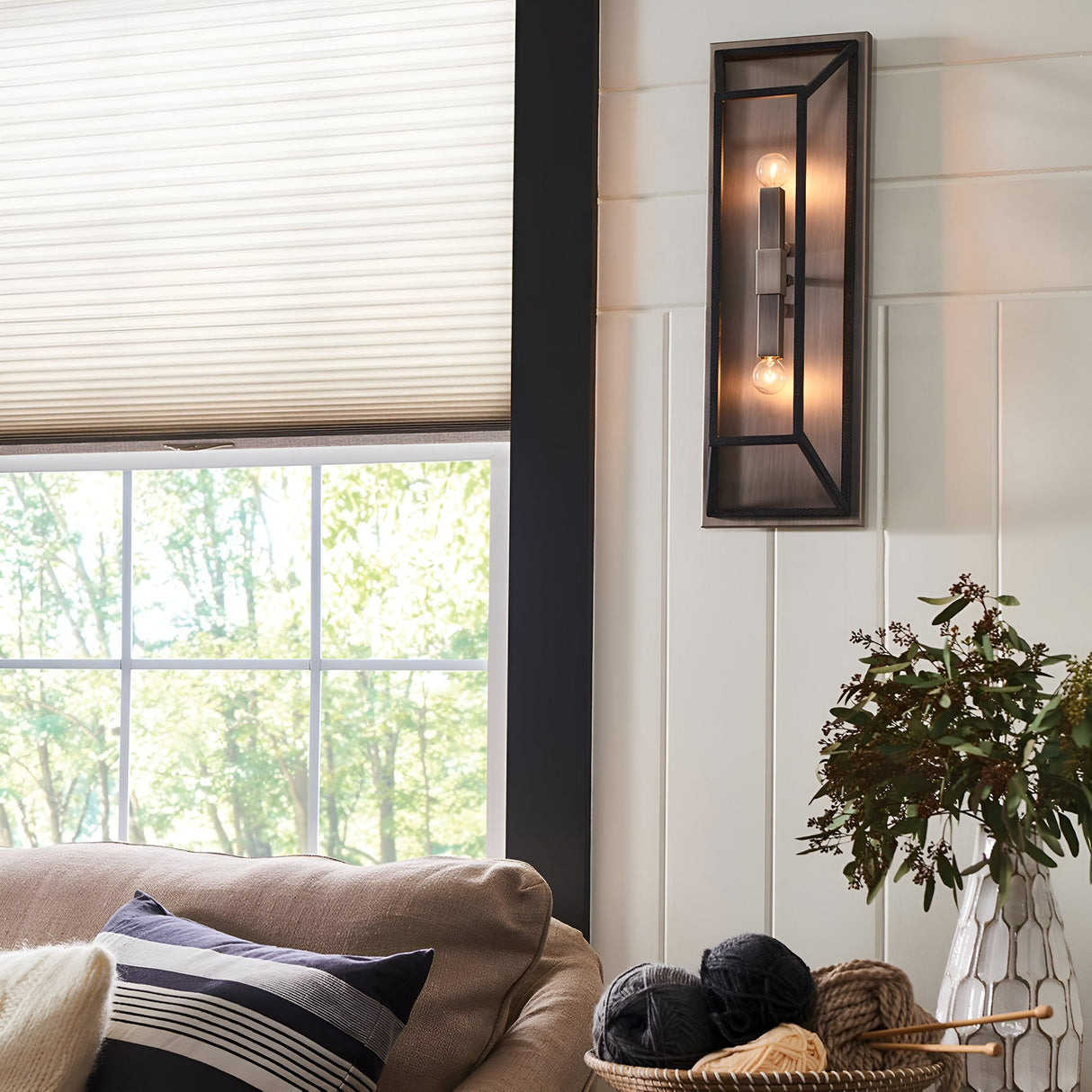 A cozy living room showcases a beige sofa with striped pillows. The space is illuminated by the Fulton 2 Light Wall Light in bronze and brass, adding a vintage industrial touch. A nearby basket with yarn complements the scene, while a window with blinds frames views of green foliage outside, embodying minimalist design elements.