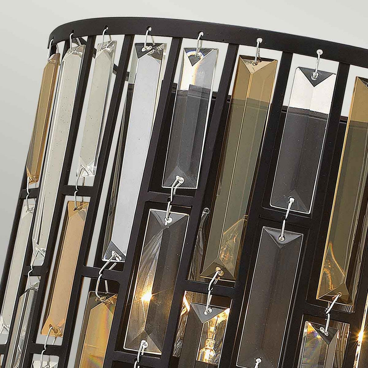 A close-up of the Gemma-1 2 Light Wall Light in Vintage Bronze showcases its stylish, modern design with rectangular glass panels in clear, beige, and smoke gray hues. The black metal frame supports an ambient glow that radiates like an amber pearl crystal, exuding contemporary sophistication.