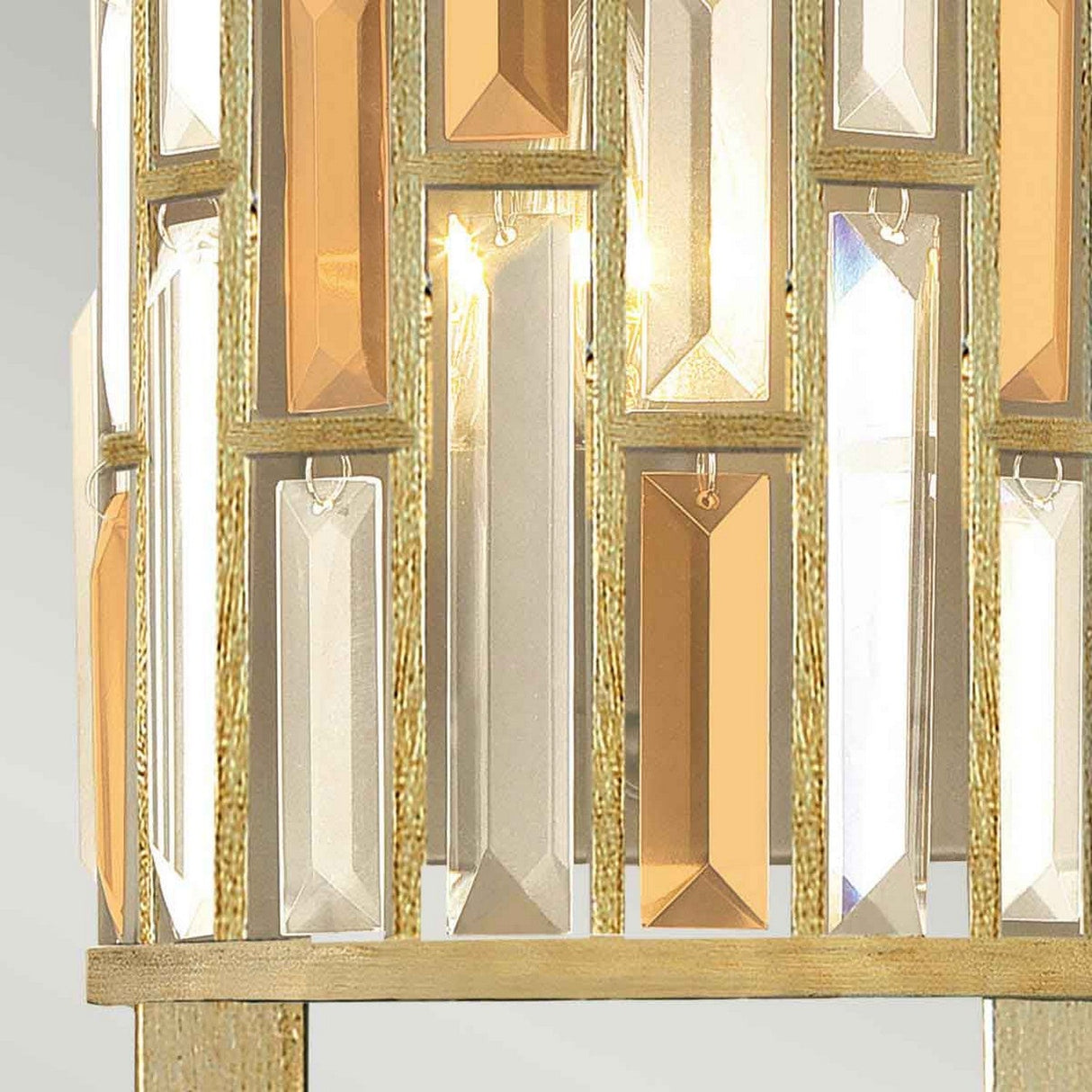 A close-up of the Gemma-2 2 Light Wall Light - Gold features a decorative fixture with rectangular, vertically arranged gold and clear glass prisms set in a gold frame. These prisms reflect and refract light, producing a shimmering effect characteristic of contemporary lighting designs.