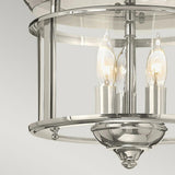 A close-up of the Gentry 3 Light Flush in polished nickel highlights its cylindrical glass shade, within which two candle-shaped bulbs emit a warm glow, offering a nod to traditional design elements. This modern chandelier contrasts beautifully against the simple light gray backdrop.