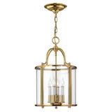 The Gentry 4 Light Medium Pendant in polished brass showcases a timeless design with clear bent glass panels that enclose four candle-shaped bulbs, delivering an elegant and classic aesthetic.
