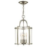 The Gentry 4 Light Medium Pendant in polished nickel hangs gracefully from a chain, showcasing a chic lantern design. Its clear glass panels encase four candle-shaped bulbs, offering an elegant and modern lighting fixture.