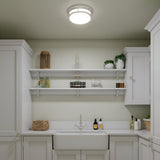A tidy, modern laundry room showcases white cabinets and a farmhouse sink. Two white shelves display decor items, plants, and laundry supplies. The space is illuminated by a Hadley 3 Light Flush - Chrome fixture with an etched opal glass shade, adding a touch of elegance to the ceiling.