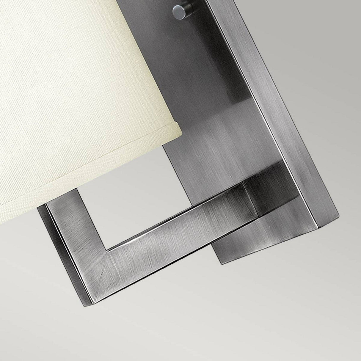 Close-up of a sleek silver wall lamp from Hampton, featuring an antique nickel rectangular metal frame and an ivory drum shade. Mounted on a gray wall, it highlights its elegant, minimalistic design accented by a decorative etched acrylic bottom lens.